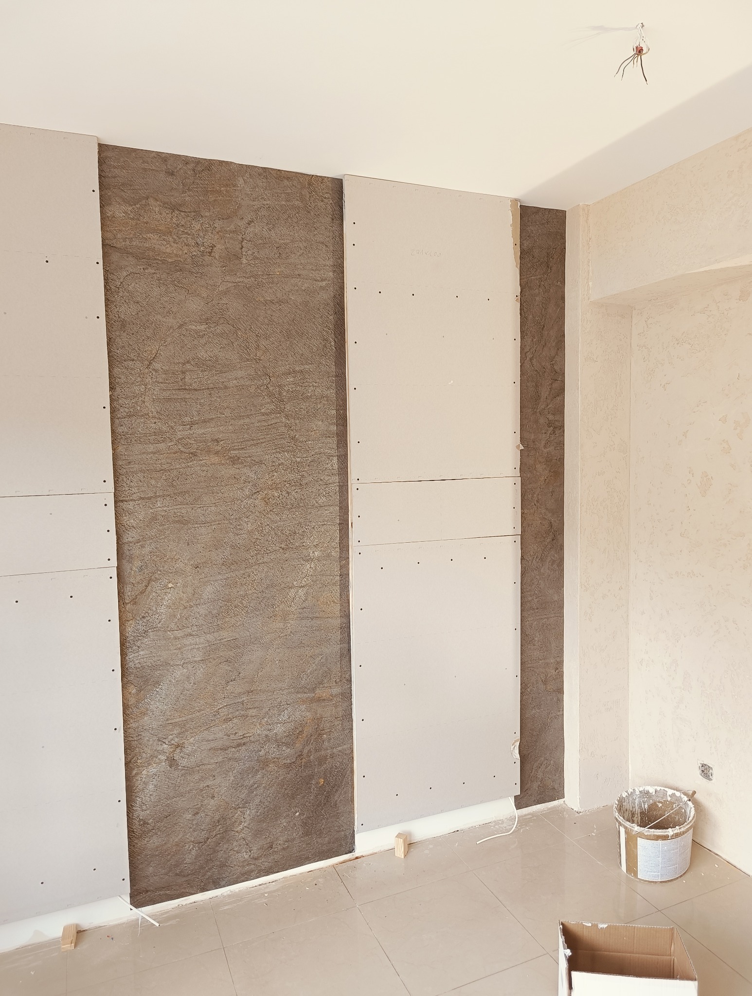 Montaż płyt gipsowo-kartonowych na ścianie z dekoracyjną okładziną. Widoczny etap prac wykończeniowych, narzędzia i materiały budowlane.