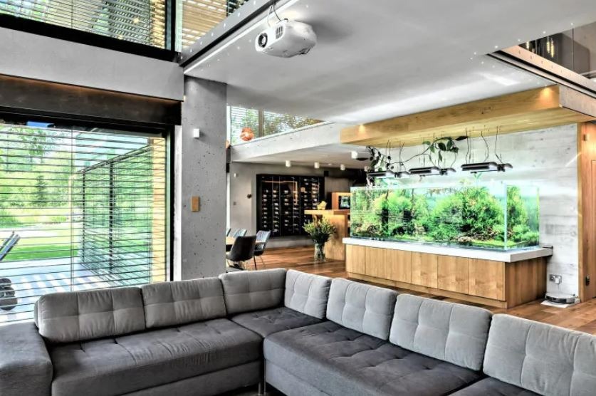 Nowoczesny salon z szarą sofą narożną, dużym akwarium z roślinami, projektorem sufitowym i widokiem na zieleń przez okno z żaluzjami.