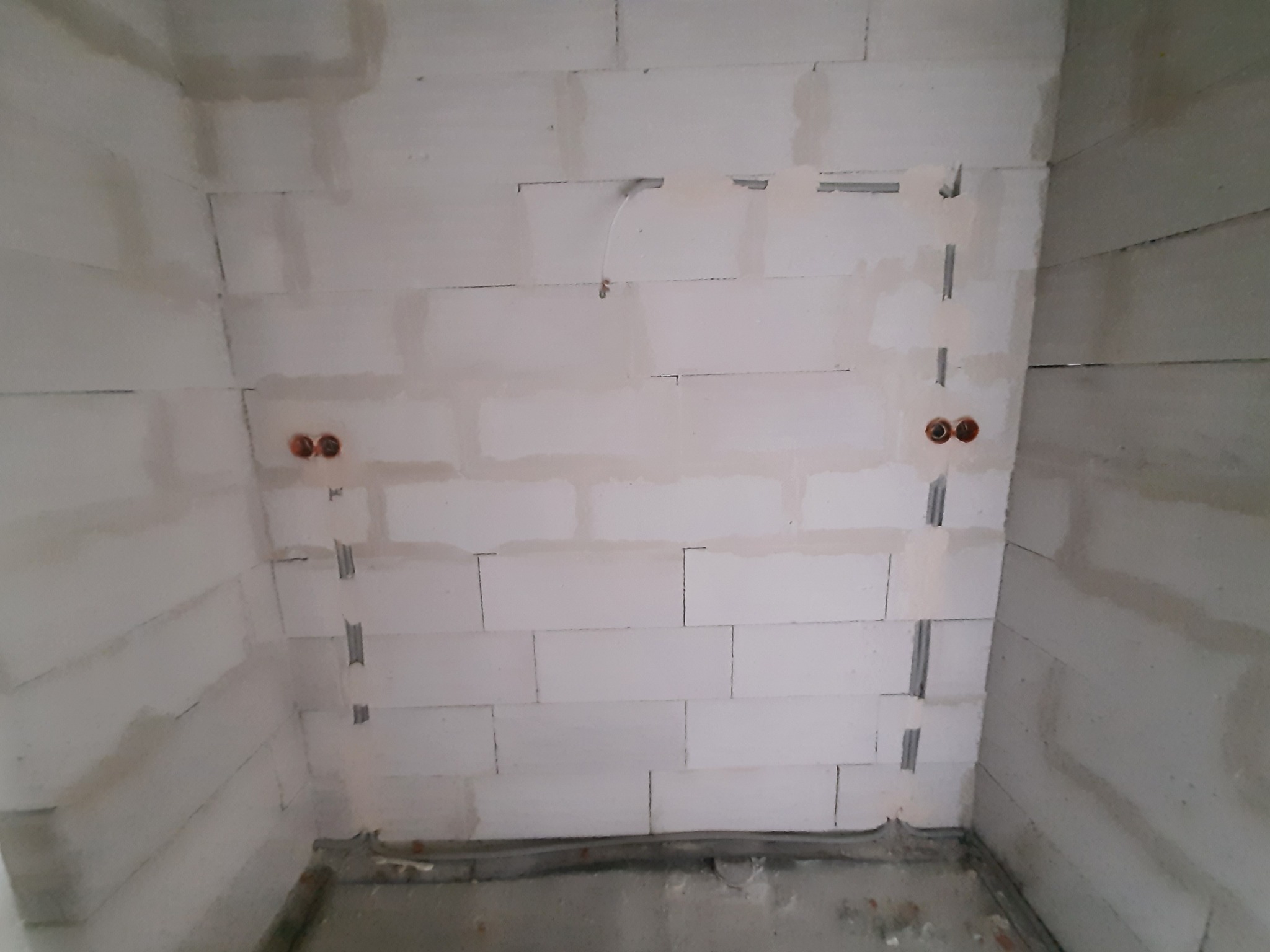 Ściana z białych bloczków, z wyciętymi bruzdami pod instalację elektryczną i widocznymi puszkami elektrycznymi, w trakcie prac budowlanych.
