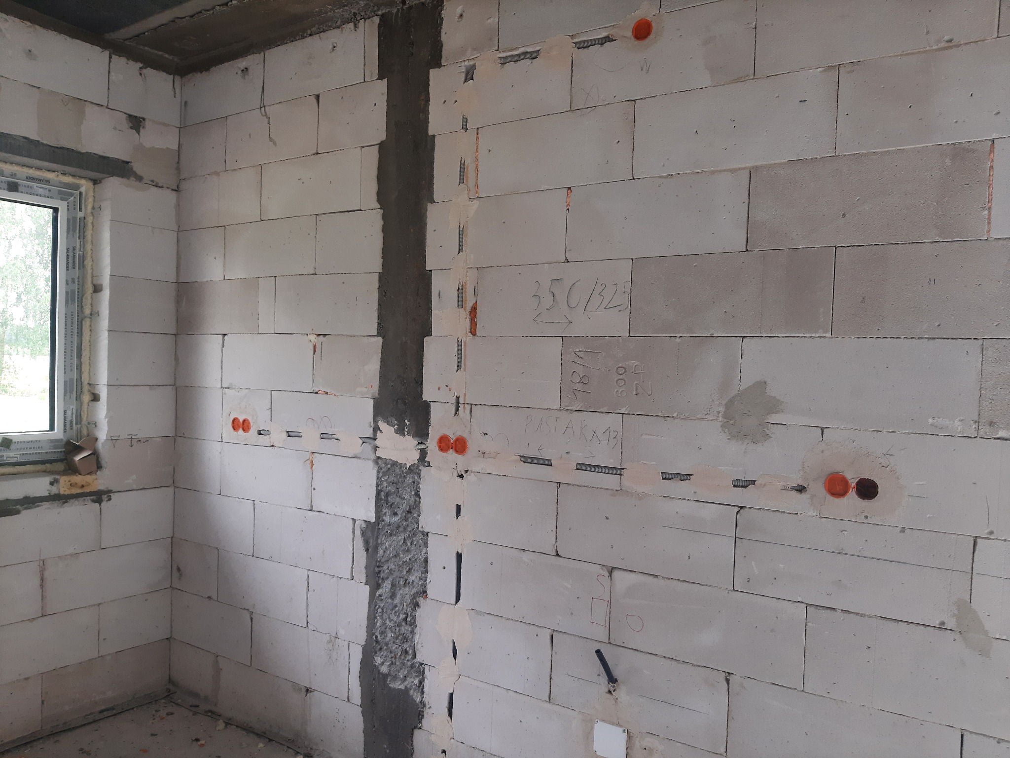 Ściana z betonu komórkowego z widocznymi puszkami instalacyjnymi w kolorze pomarańczowym, metalowymi peszlami i pionową bruzdą wypełnioną zaprawą. Na ścianie widoczne odręczne notatki i oznaczenia.