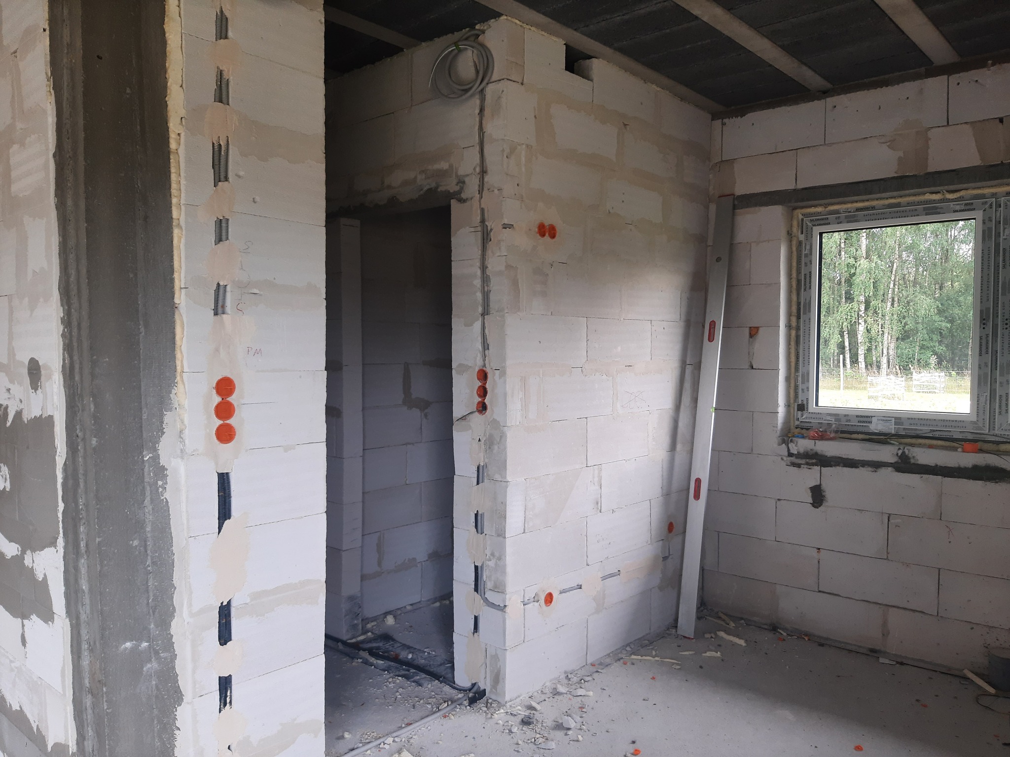 Wnętrze w stanie surowym: ściany z bloczków, widoczne przygotowane punkty elektryczne, w podłodze poprowadzone kable, okno z widokiem na zewnątrz, poziomica oparta o ścianę.