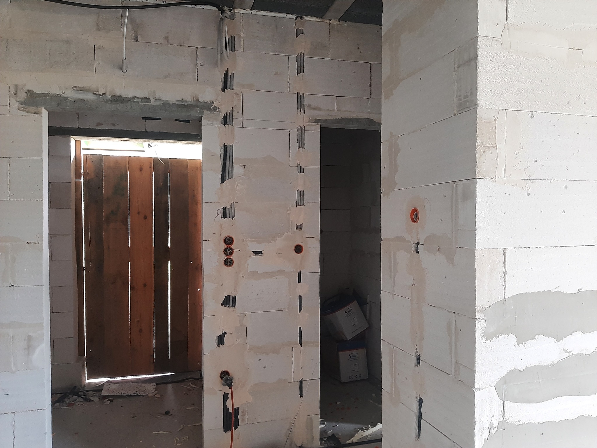 Wnętrze w trakcie budowy z białych bloków, widoczne bruzdy pod instalacje elektryczne i otwory pod puszki, niezakończone ościeże drzwiowe z widoczną drewnianą konstrukcją w tle.