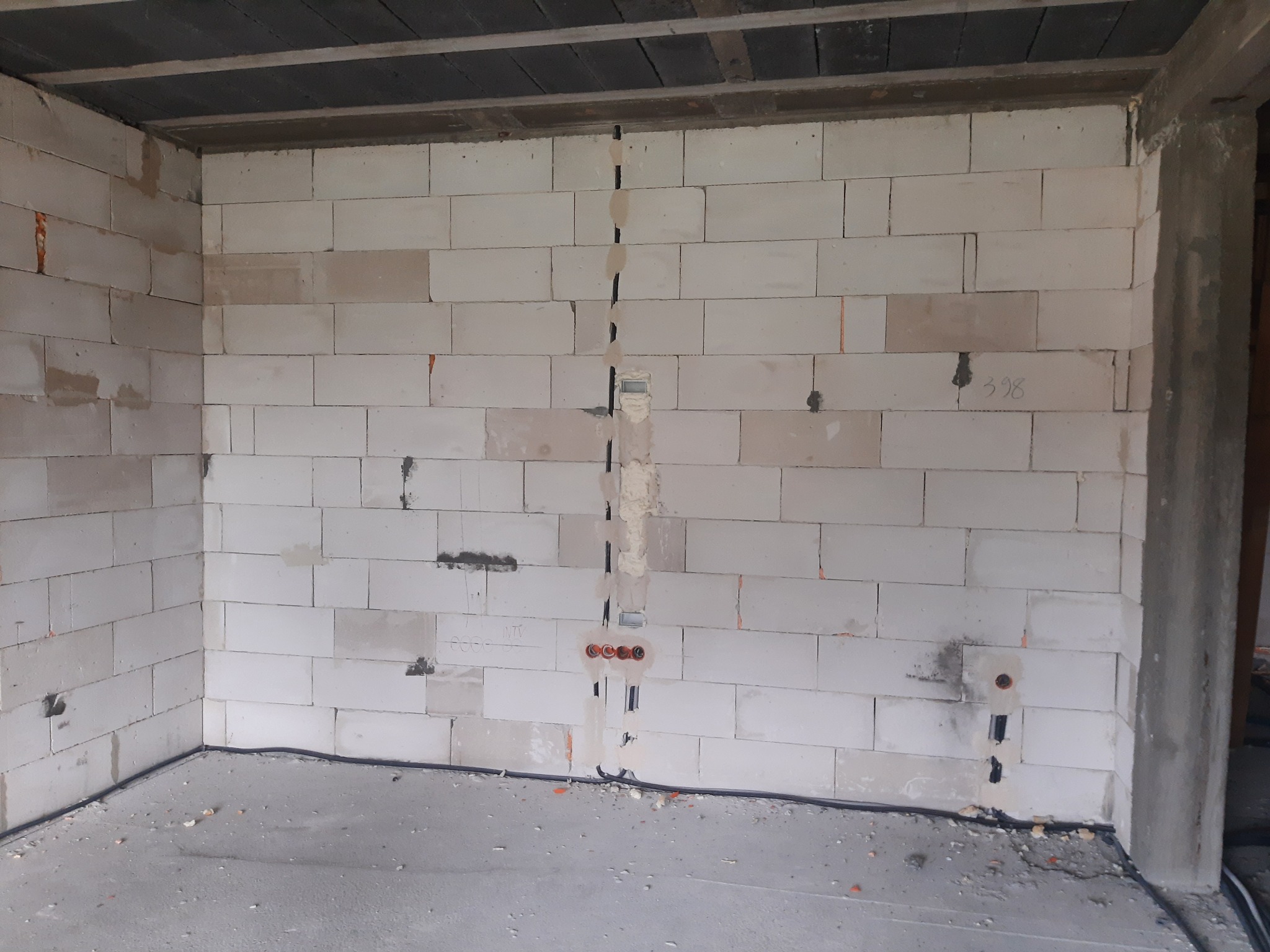 Wnętrze w trakcie budowy: ściany z białych bloczków, pionowy kanał instalacyjny z przewodami elektrycznymi i puszkami, surowy beton na podłodze i suficie.