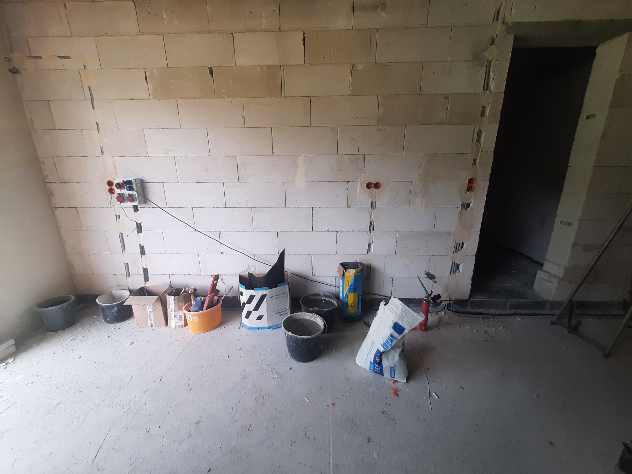 Surowy stan wnętrza z widocznymi instalacjami elektrycznymi w bruzdach, pustaki, worki z zaprawą, wiadra i narzędzia przygotowane do dalszych prac wykończeniowych.
