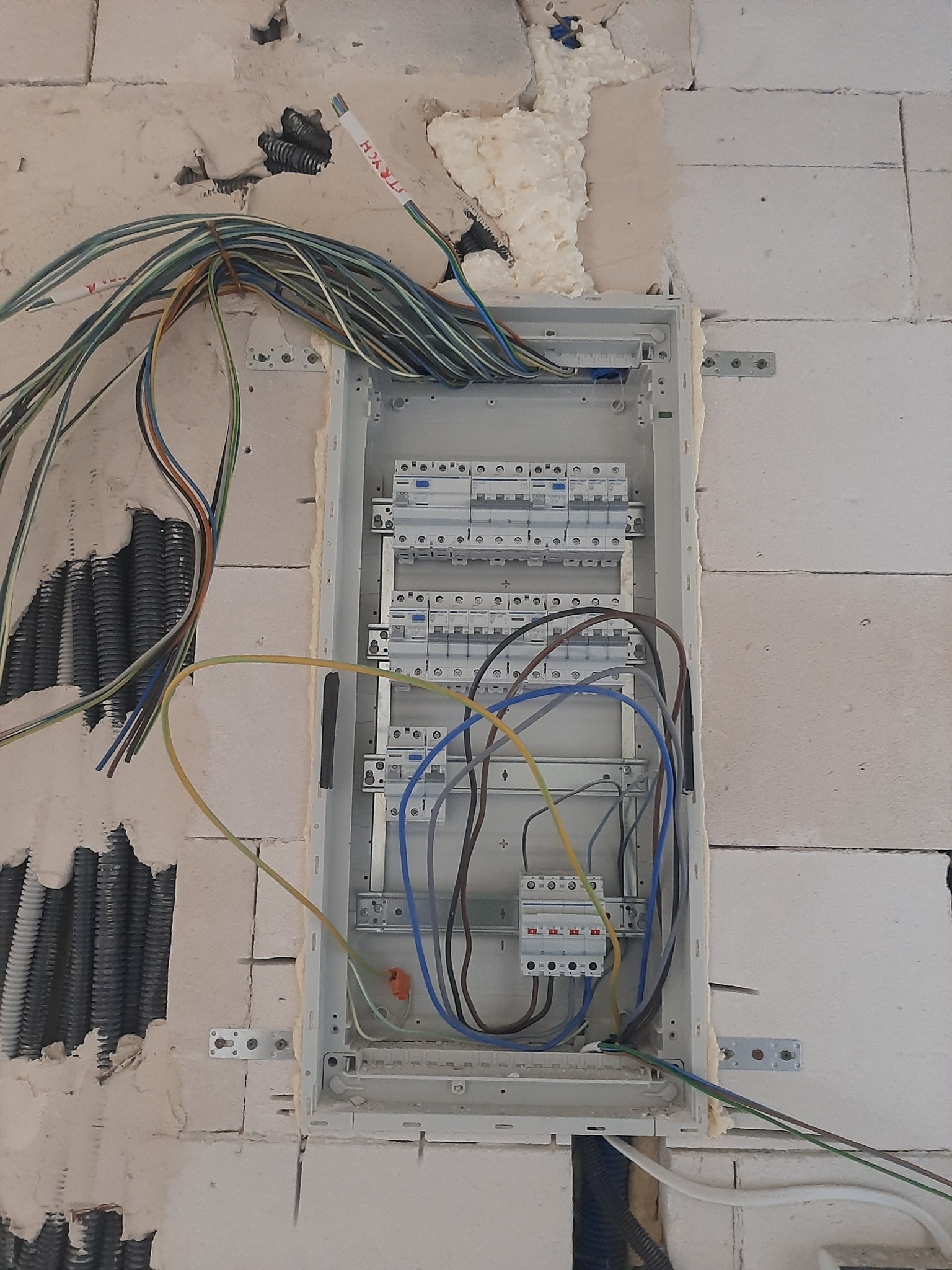 Rozdzielnica elektryczna w trakcie montażu w ścianie z betonu komórkowego, widoczne przewody, peszle i pianka montażowa.