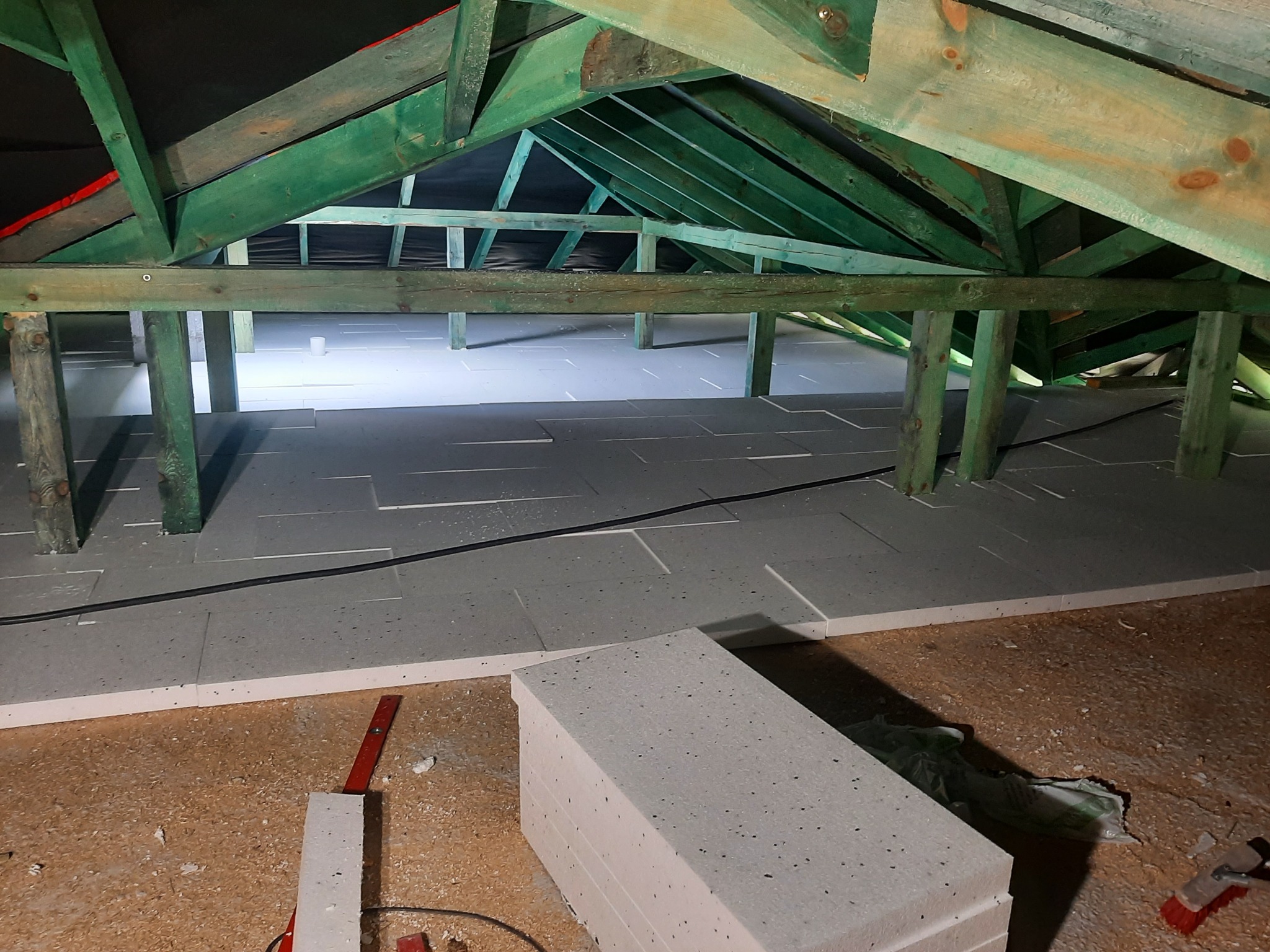 Ocieplenie poddasza wełną mineralną lub styropianem, widoczna konstrukcja drewniana dachu pomalowana na zielono i ułożone płyty izolacyjne z widocznymi drobnymi czarnymi plamkami.