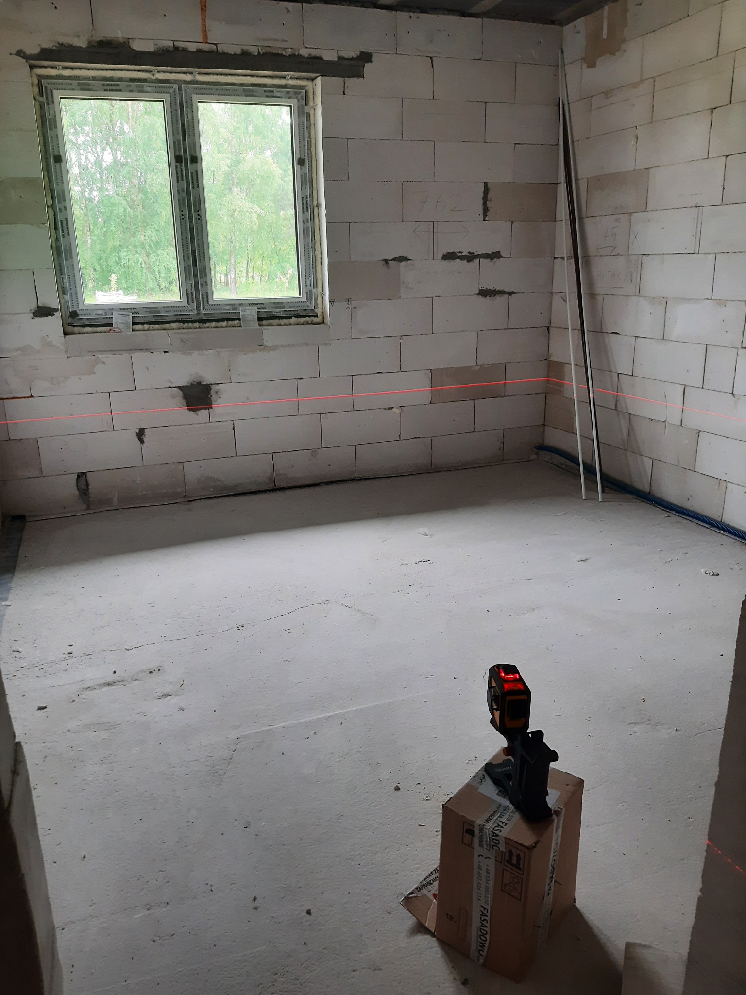 Surowy pokój z oknem w trakcie prac wykończeniowych, widoczne ściany z betonu komórkowego, w tle widok zieleni za oknem, na pierwszym planie niwelator laserowy ustawiony na kartonie, wyznaczający...