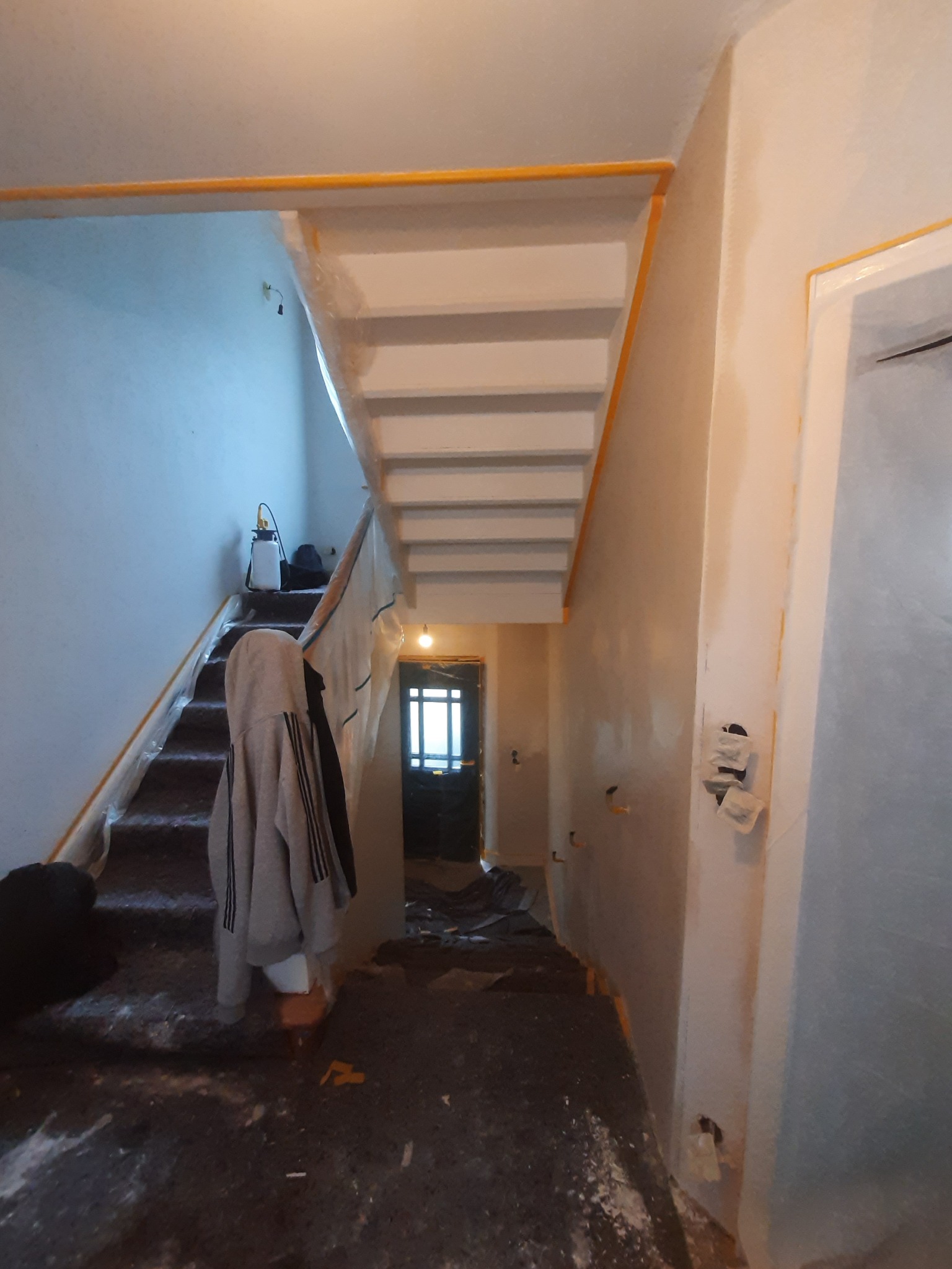 Klatka schodowa w trakcie prac przygotowawczych do malowania, zabezpieczona folią i taśmą malarską, widoczne ślady po szpachlowaniu na ścianach, podłoga zabezpieczona folią malarską.