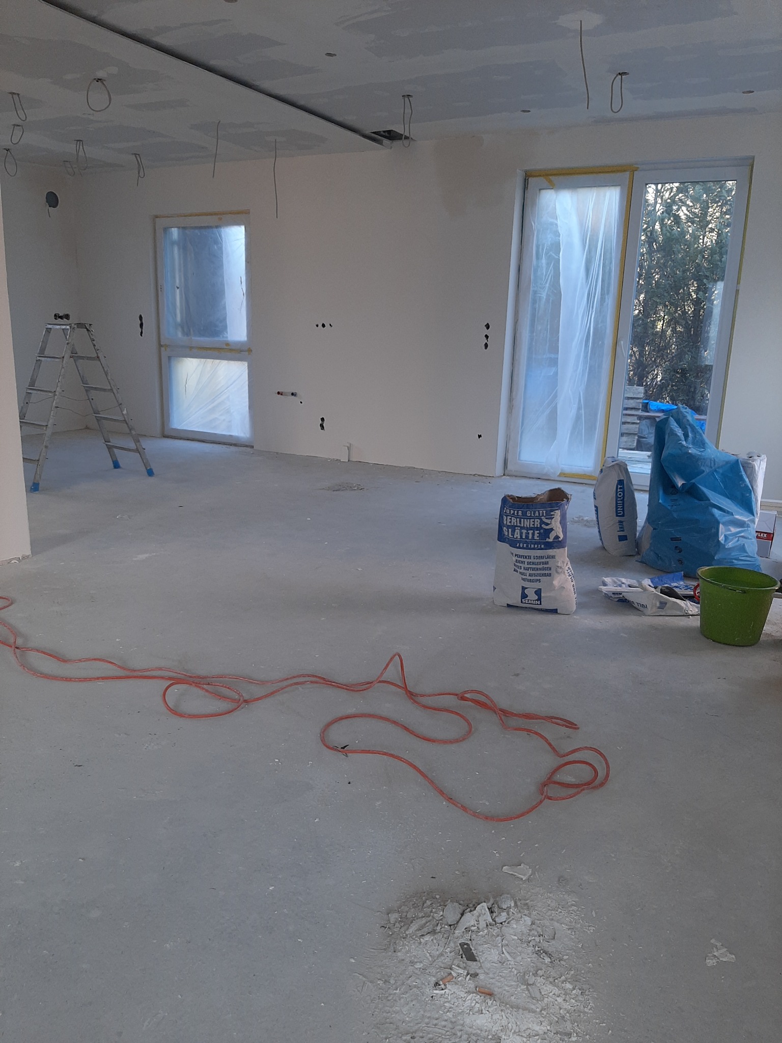 Pokój w trakcie remontu, widoczne worki z gładzią szpachlową, drabina, przewody elektryczne zwisające z sufitu oraz okna zabezpieczone folią malarską.