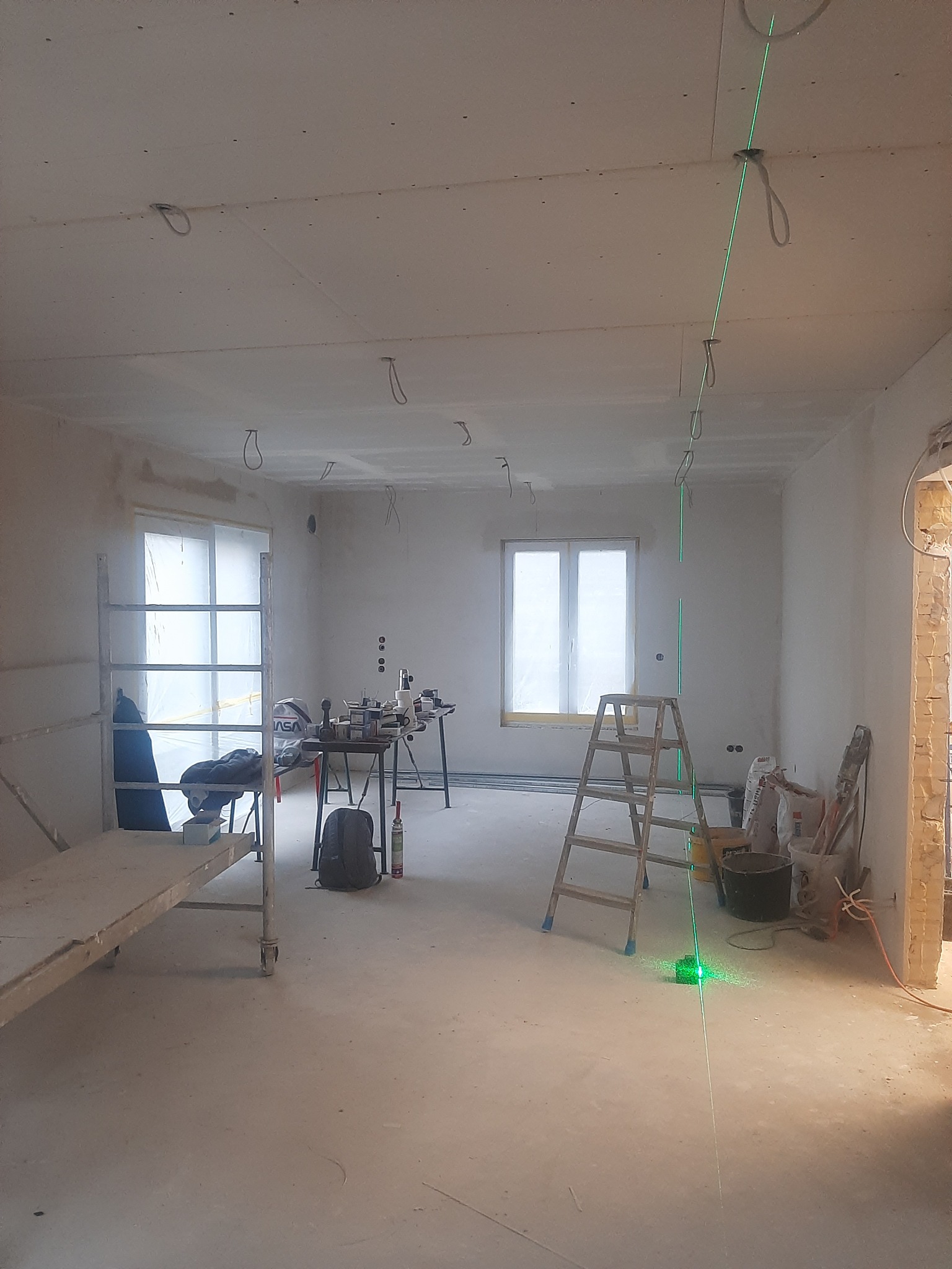 Pokój w trakcie prac wykończeniowych: ściany i sufit pokryte płytami gipsowo-kartonowymi, widoczny laser krzyżowy wyznaczający pion, drabina, rusztowanie, narzędzia i materiały budowlane.