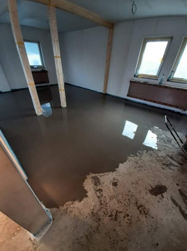 Wylewka samopoziomująca w pokoju z dwoma oknami, drewnianymi belkami i słupami zabezpieczonymi folią, częściowo jeszcze nie rozprowadzona.