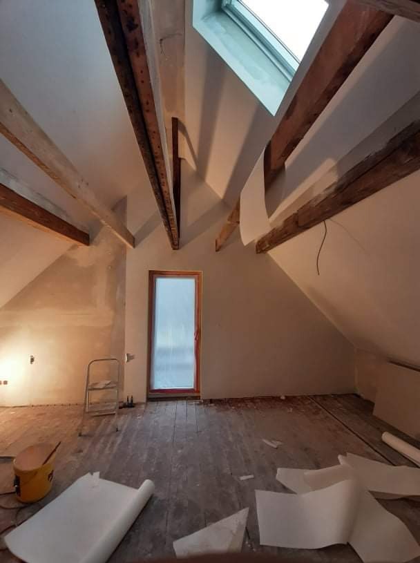 Poddasze w trakcie prac, widoczne drewniane belki stropowe, okno dachowe, przygotowane ściany do malowania oraz rozłożona folia ochronna na drewnianej podłodze, drabina oparta o ścianę.