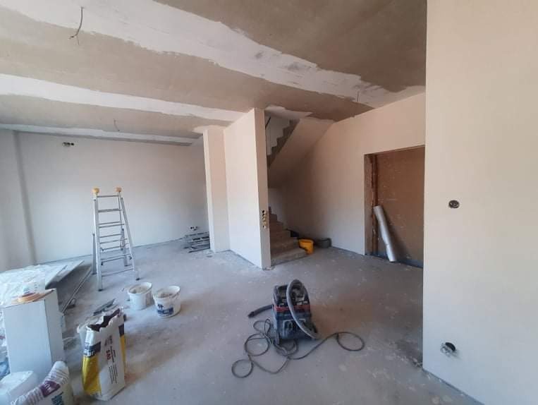 Wnętrze w trakcie prac wykończeniowych, widoczne świeżo pomalowane ściany, surowy sufit z widocznymi belkami, drabina aluminiowa, worki z materiałami budowlanymi i odkurzacz przemysłowy...