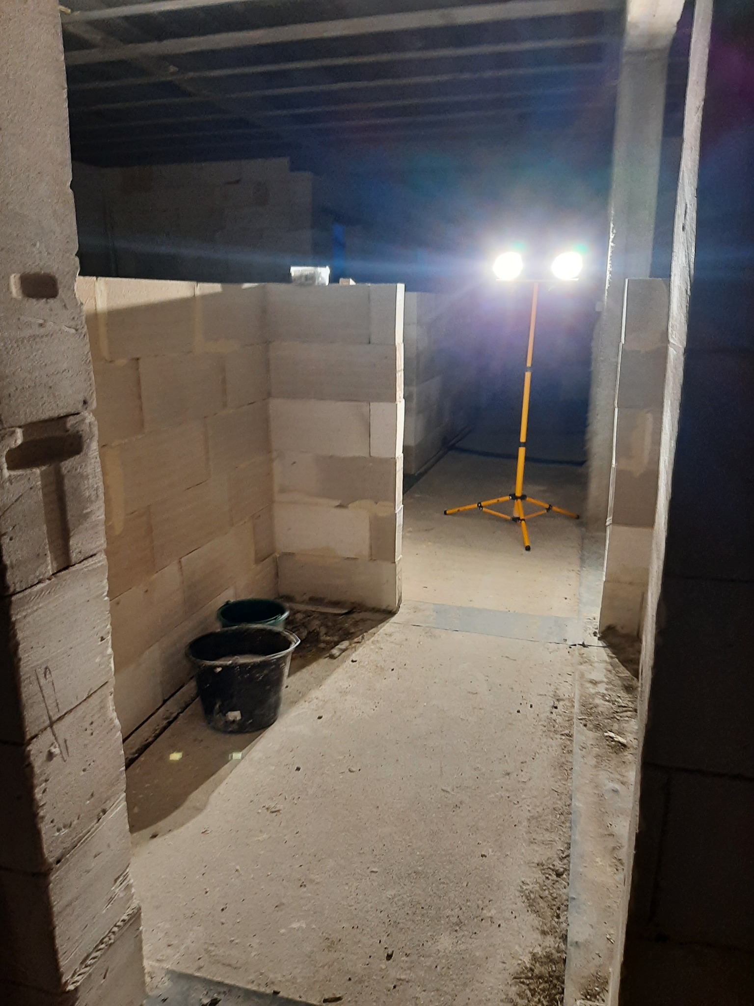 Wnętrze w trakcie budowy z białych bloczków, oświetlone lampą budowlaną na żółtym statywie, widoczne dwa plastikowe wiadra.