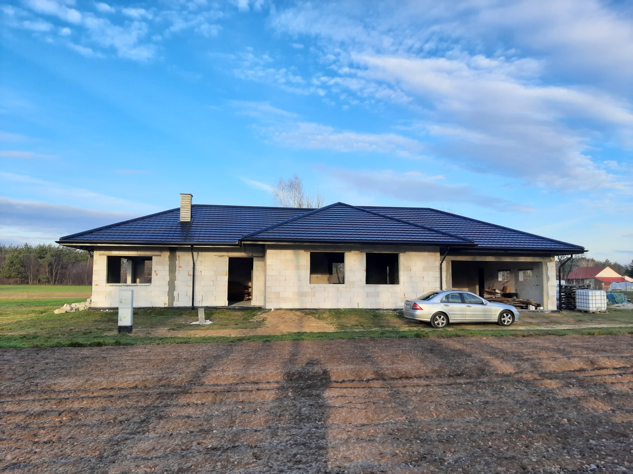 Niedokończony dom jednorodzinny z białego bloczka, z ciemną dachówką, widoczny samochód osobowy na tle nieba z chmurami.