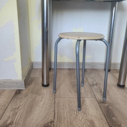 Rafał Stolarczyk - Fragment pokoju z podłogą z paneli, przy ścianie z widocznymi śladami po starej farbie, na podłodze stoi metalowy stołek pod stołem.