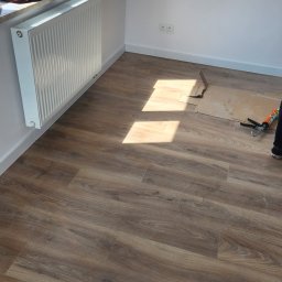 Rafał Stolarczyk - Układanie paneli podłogowych w jasnym pokoju, widoczne narzędzia i fragment kartonu ochronnego, naturalne światło podkreśla strukturę drewna.