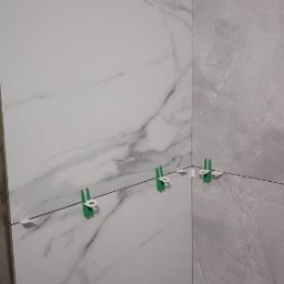 Rafał Stolarczyk - Montaż płytek imitujących marmur w rogu łazienki, widoczne zielone kliny poziomujące i żyłka dystansowa.