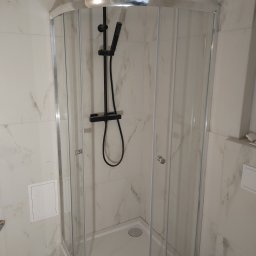 Rafał Stolarczyk - Kabina prysznicowa narożna z czarną armaturą, białe płytki imitujące marmur, szara podłoga, widok na wnętrze kabiny.