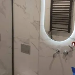 Nowoczesna łazienka z marmurowymi płytkami, podwieszaną toaletą i owalnym lustrem z podświetleniem LED, w którym odbija się grzejnik drabinkowy.