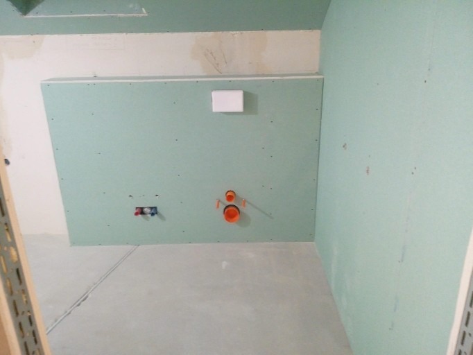 Ściana w trakcie przygotowywania do montażu podwieszanej toalety, widoczne instalacje wodno-kanalizacyjne i stelaż obudowany płytami gipsowo-kartonowymi, przygotowany do dalszych prac wykończeniowych.