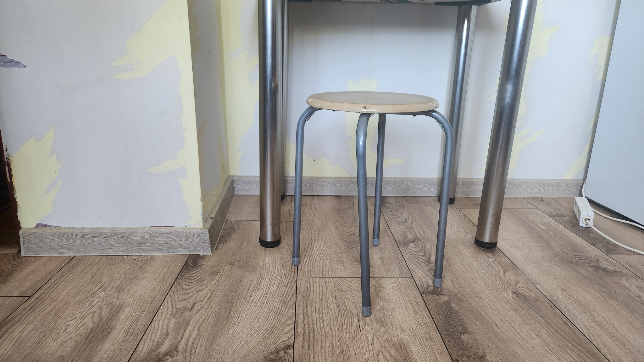 Fragment pokoju z podłogą z paneli, przy ścianie z widocznymi śladami po starej farbie, na podłodze stoi metalowy stołek pod stołem.