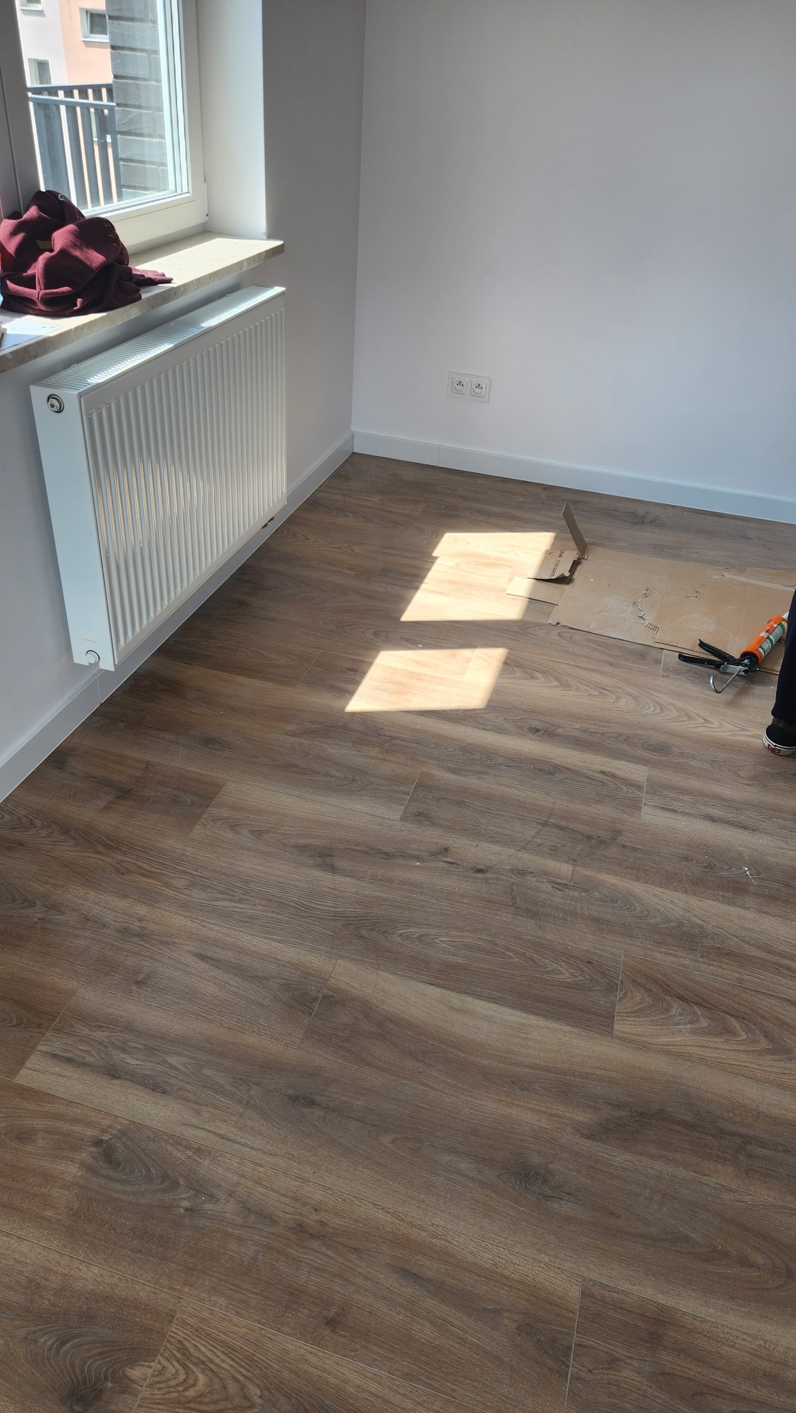 Układanie paneli podłogowych w jasnym pokoju, widoczne narzędzia i fragment kartonu ochronnego, naturalne światło podkreśla strukturę drewna.