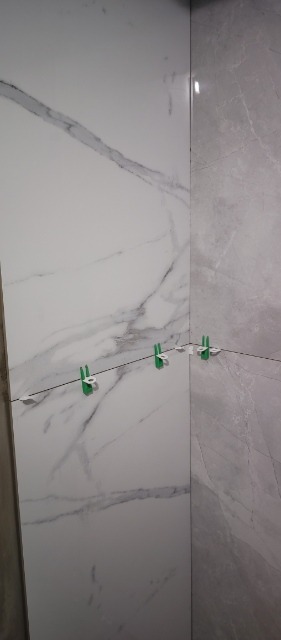 Montaż płytek imitujących marmur w rogu łazienki, widoczne zielone kliny poziomujące i żyłka dystansowa.