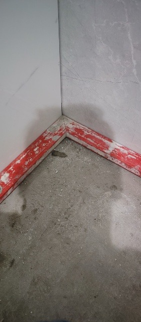 Fragment narożnika w trakcie zabezpieczania folią malarską przed pracami wykończeniowymi, widoczne dwie ściany wyłożone płytkami w różnych odcieniach szarości i surowa betonowa podłoga.