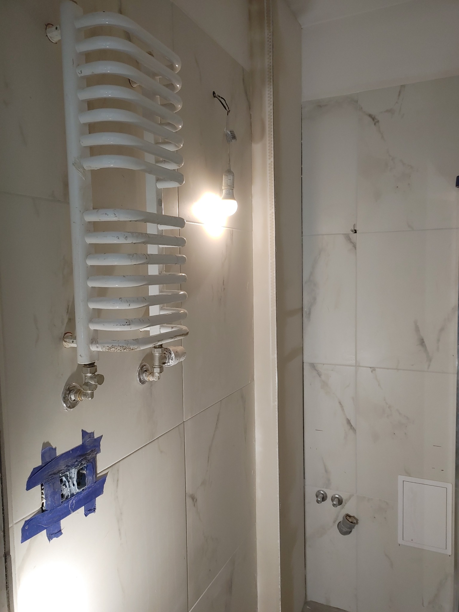 Łazienka w trakcie remontu: białe marmurowe płytki, grzejnik drabinkowy z widocznymi śladami użytkowania, prowizoryczne oświetlenie żarówką, przygotowane miejsce pod gniazdko zabezpieczone taśmą.