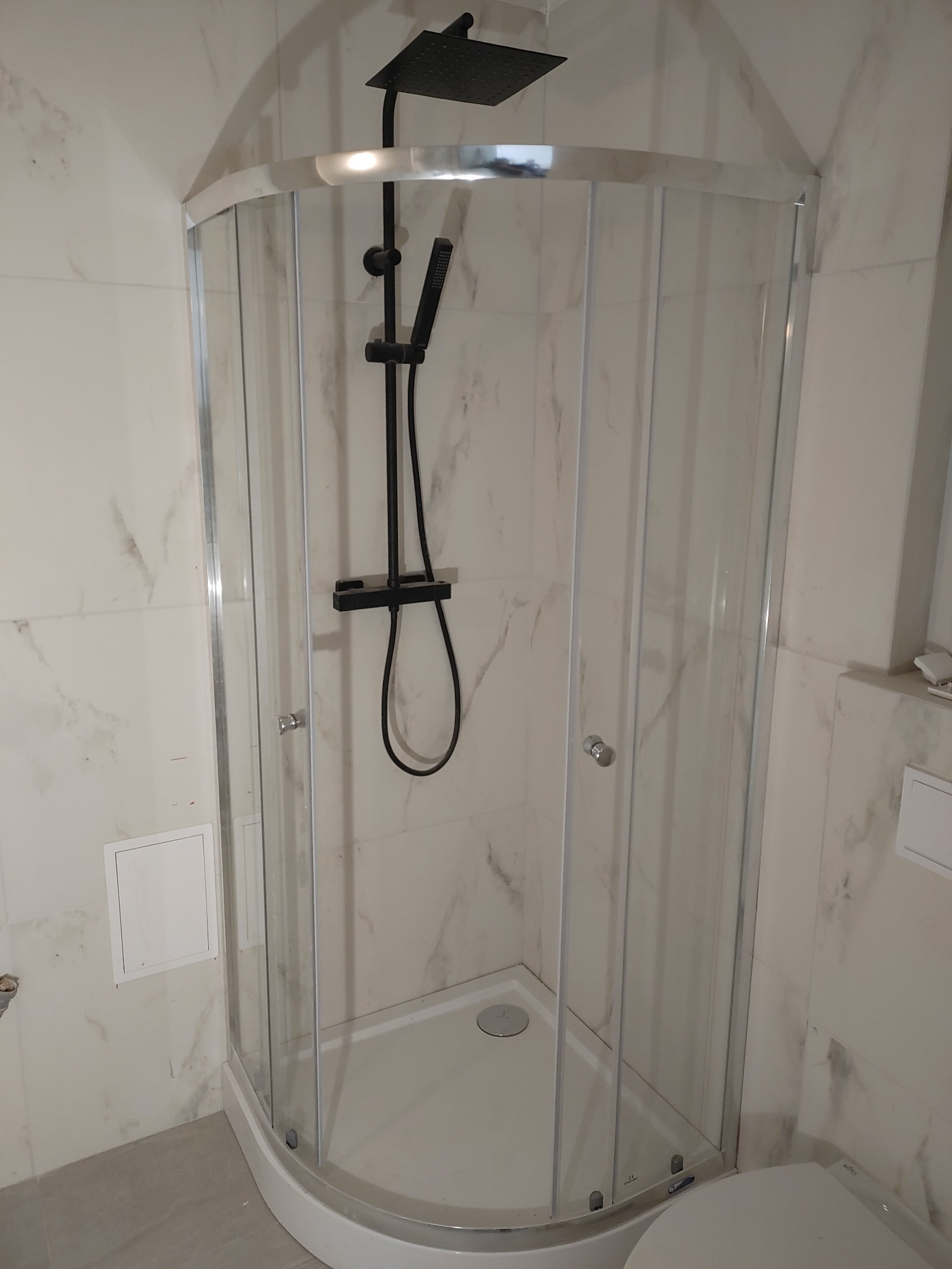 Kabina prysznicowa narożna z czarną armaturą, białe płytki imitujące marmur, szara podłoga, widok na wnętrze kabiny.