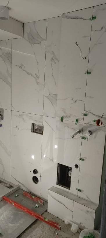 Ściana w trakcie wykańczania z białych płytek imitujących marmur, widoczne otwory na instalacje, poziomice i zielone klipsy do poziomowania płytek.