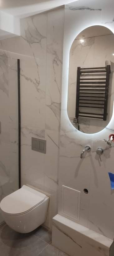 Nowoczesna łazienka z marmurowymi płytkami, podwieszaną toaletą i owalnym lustrem z podświetleniem LED, w którym odbija się grzejnik drabinkowy.