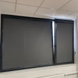 Ciemnoszare rolety zwinięte w czarnych ramach okiennych w biurze, widoczny fragment grzejnika i blatu biurka.