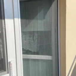 Zewnętrzna, plisowana moskitiera okienna w białej ramie, widoczny termometr okienny wskazujący temperaturę otoczenia.