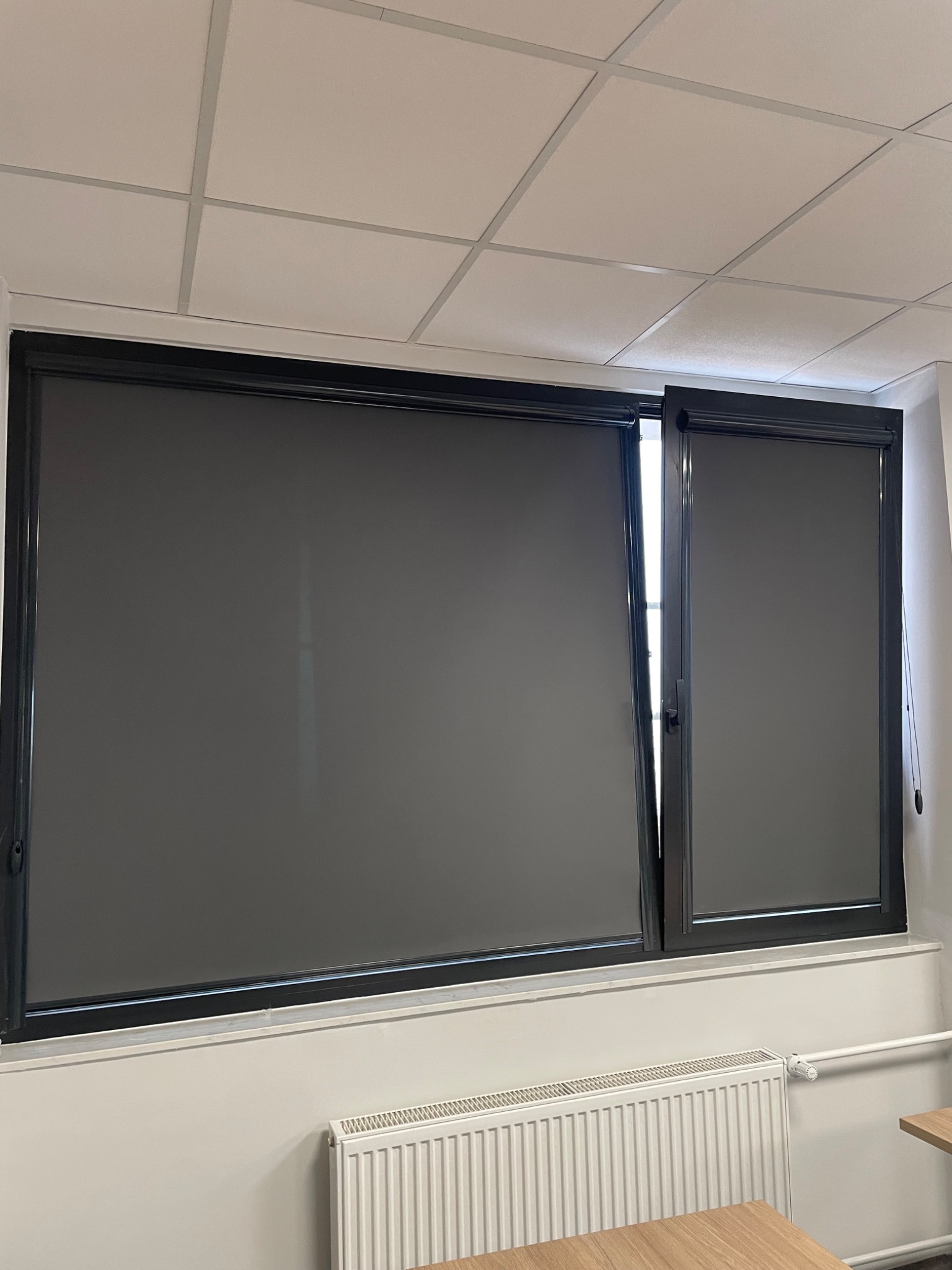 Ciemnoszare rolety zwinięte w czarnych ramach okiennych w biurze, widoczny fragment grzejnika i blatu biurka.