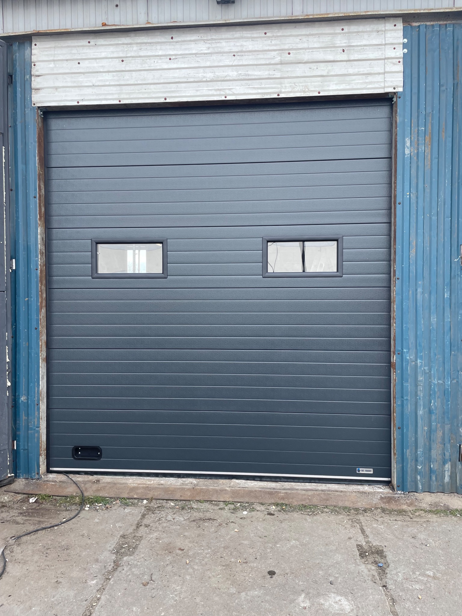 Szara segmentowa brama garażowa z dwoma małymi oknami, widoczna z zewnątrz budynku o niebieskiej, pofalowanej elewacji.