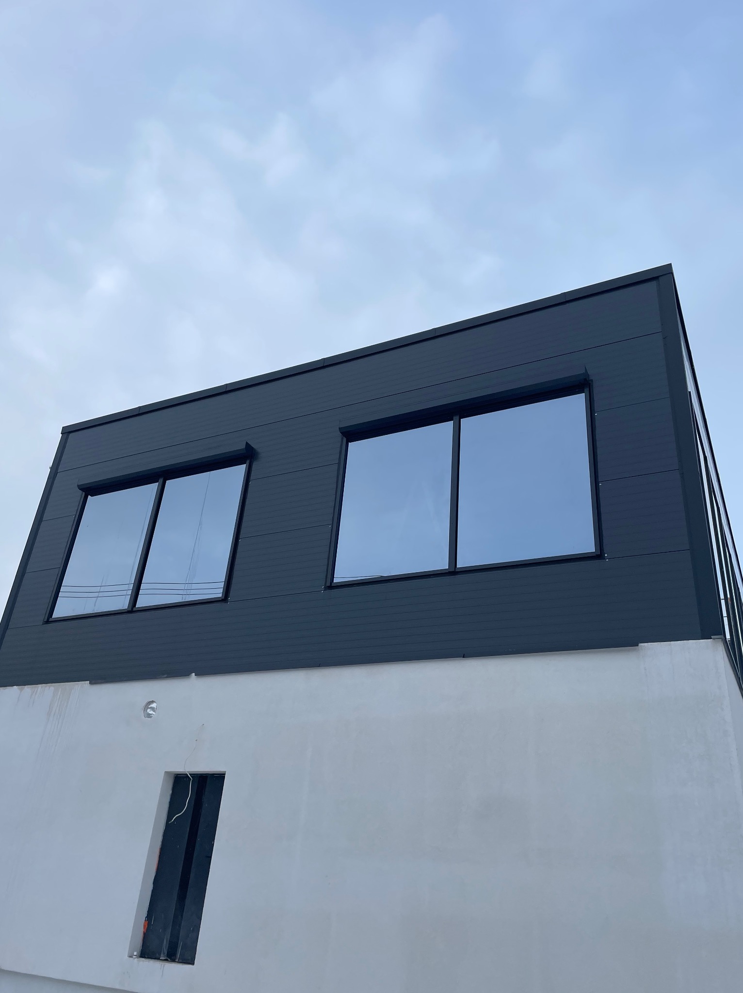 Nowoczesny budynek z szarą elewacją i dwoma oknami z zamontowanymi zewnętrznymi osłonami przeciwsłonecznymi, widok z dołu na tle pochmurnego nieba.
