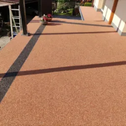 Wykończony balkon z żywicznej, pomarańczowo-brązowej posadzki z czarnym, dekoracyjnym pasem, donice z kwiatami na skraju, widoczne elementy konstrukcji dachu i drabina.