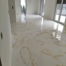 Biała, błyszcząca podłoga z efektem marmuru w przestronnym, jasnym pomieszczeniu z trzema oknami, widoczne ślady brązowego wzoru.