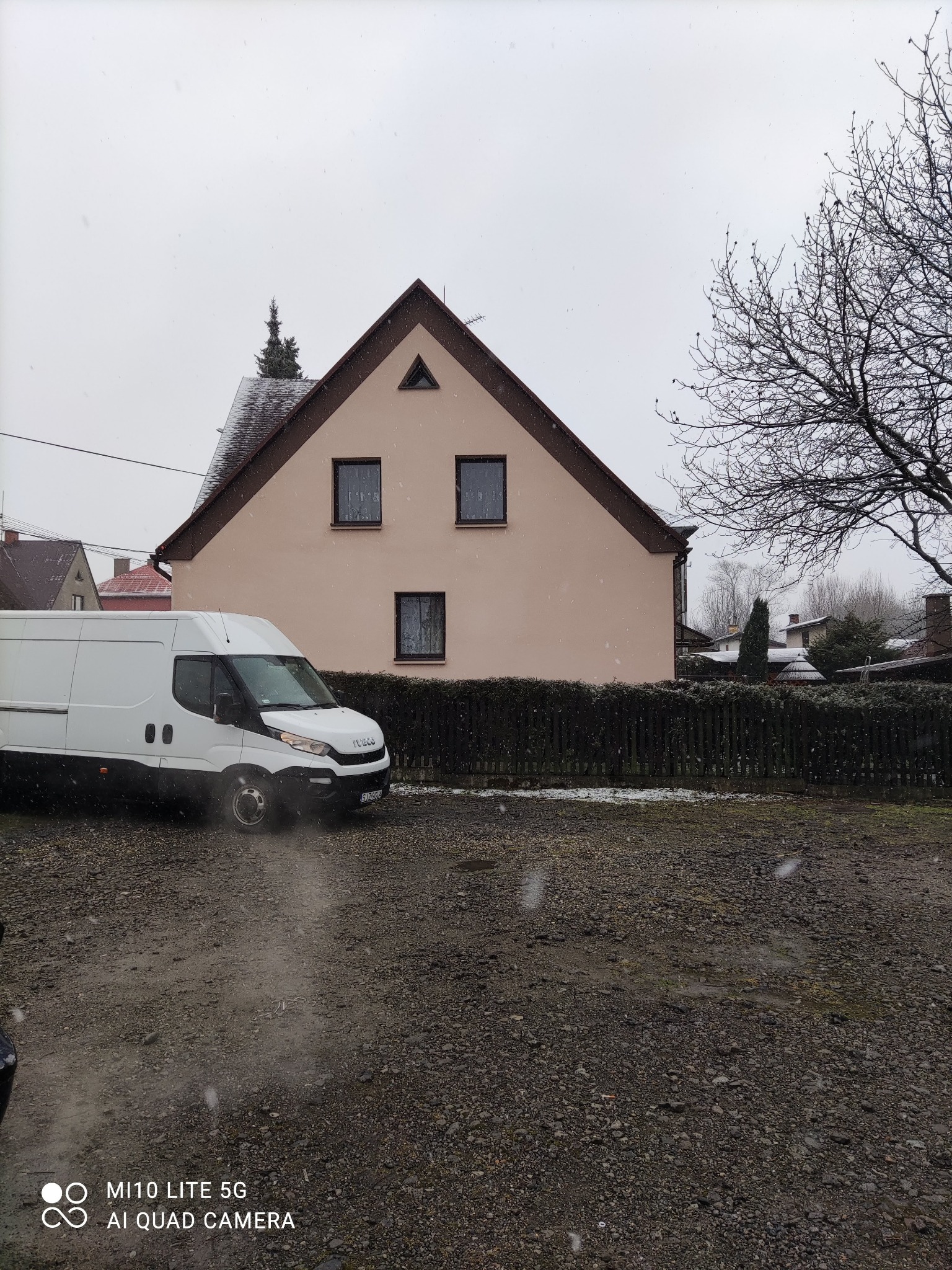 Biały samochód dostawczy Iveco zaparkowany przed domem w trakcie opadów śniegu, widoczny fragment ogrodzenia i drzewo bez liści.