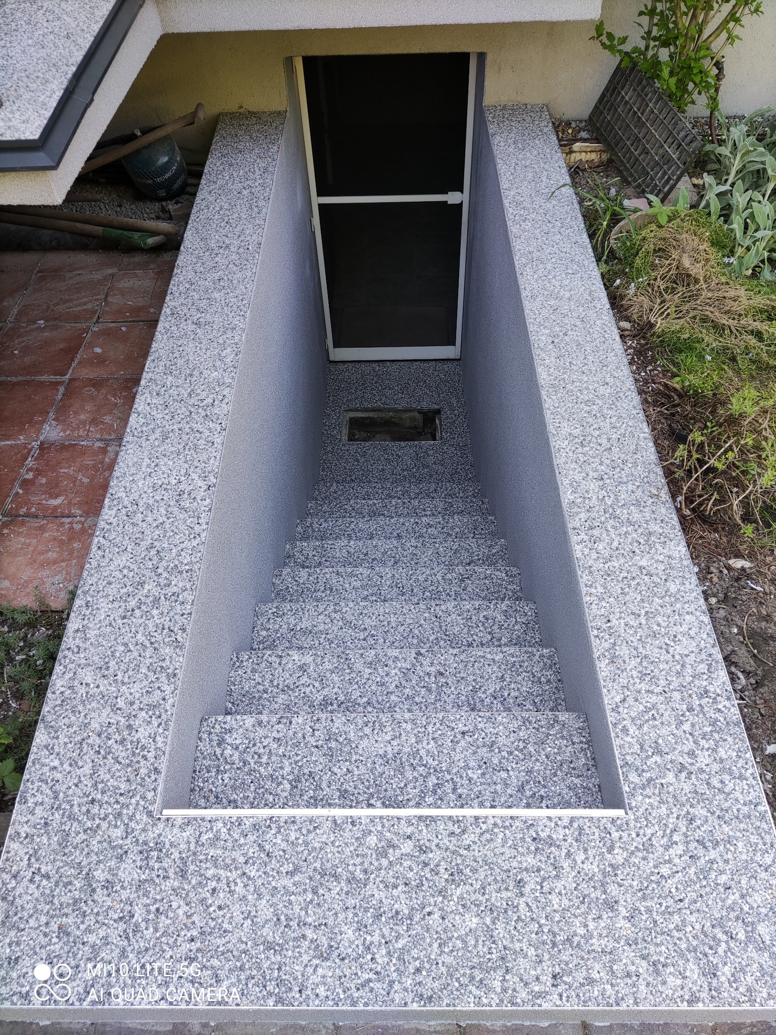 Zewnętrzne schody prowadzące do piwnicy, wykończone szarym, nakrapianym kamieniem, z metalowymi listwami na krawędziach stopni i czarnymi drzwiami na końcu schodów.