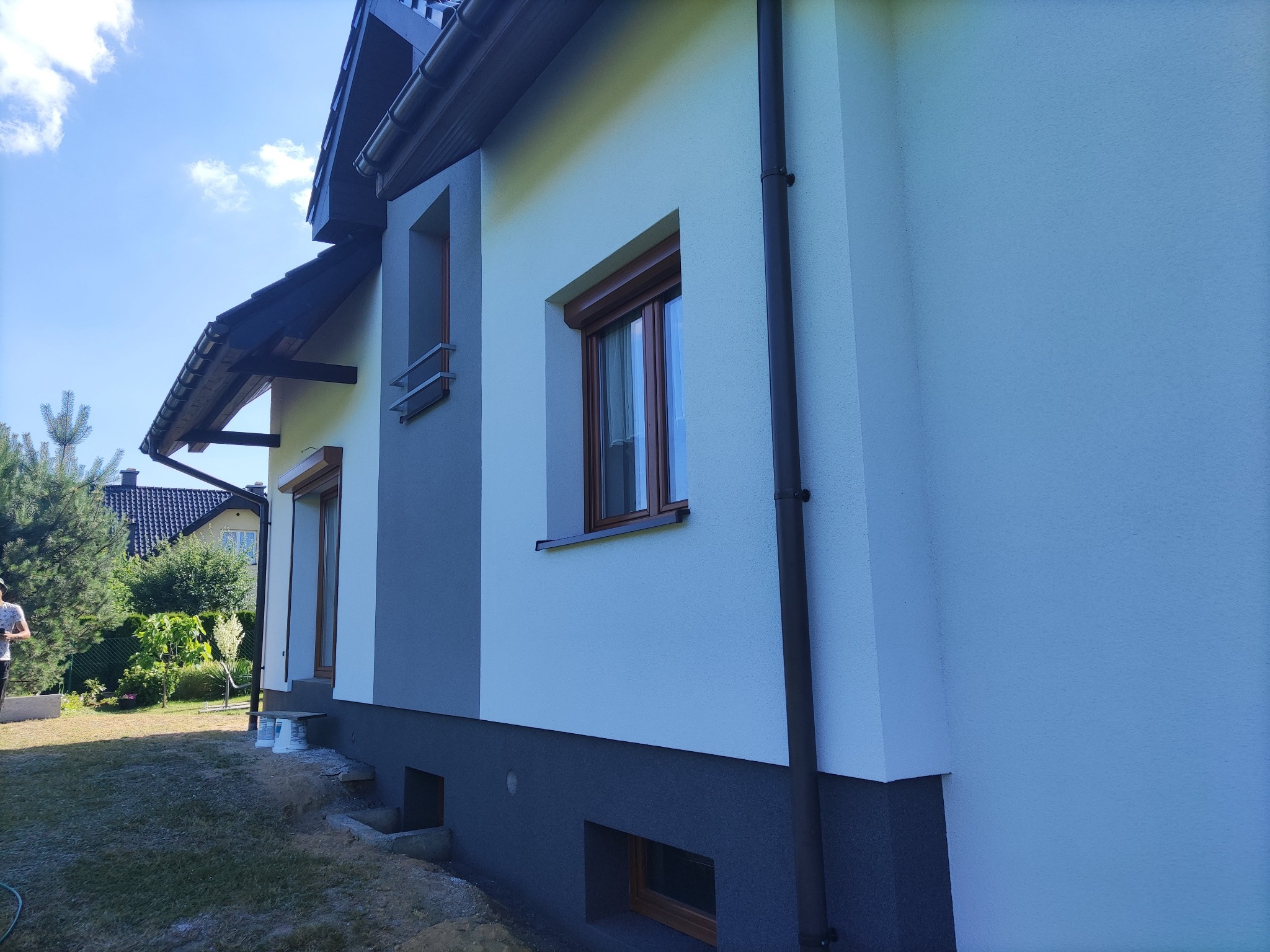 Elewacja domu jednorodzinnego po remoncie, z widocznym podziałem na szary cokół, białą górną część i szare wstawki, brązowe okna z roletami zewnętrznymi, rynna w kolorze grafitowym.