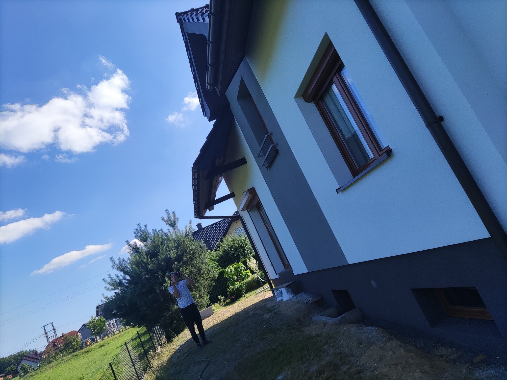 Dwukolorowa elewacja domu jednorodzinnego z drewnianą stolarką okienną, widoczna osoba robiąca zdjęcie telefonem na tle błękitnego nieba i zieleni ogrodu.