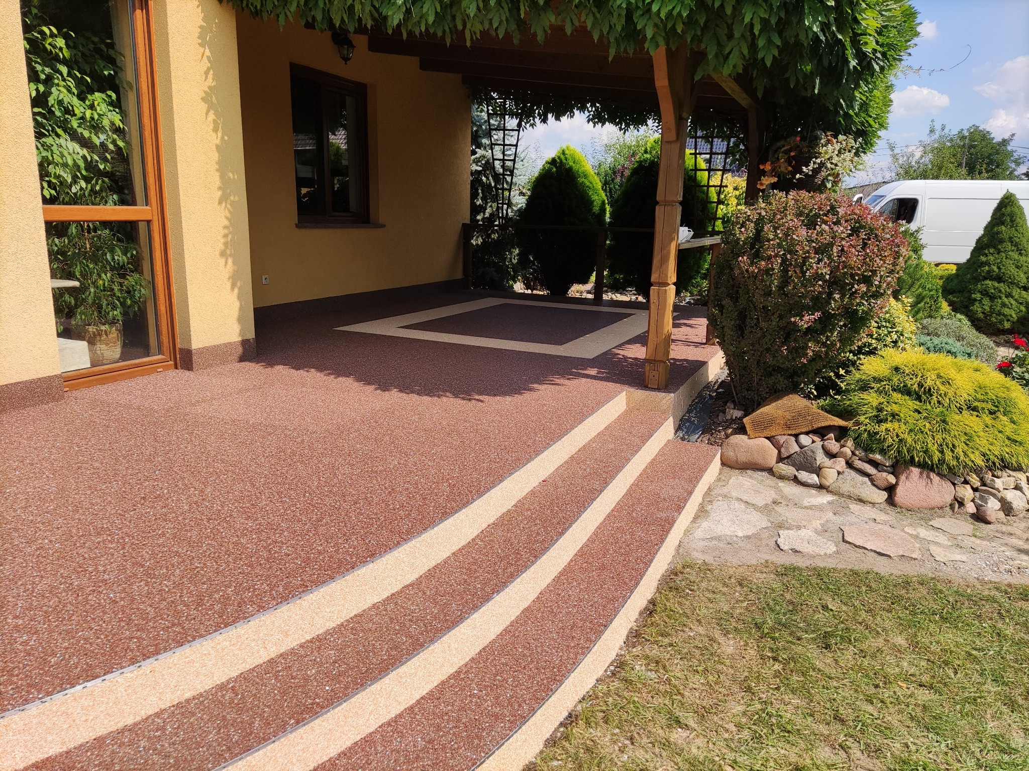 Balkon z zadaszeniem i schodami wykończony żywicą w odcieniach brązu i beżu, widok z zewnątrz na zadbany ogród.