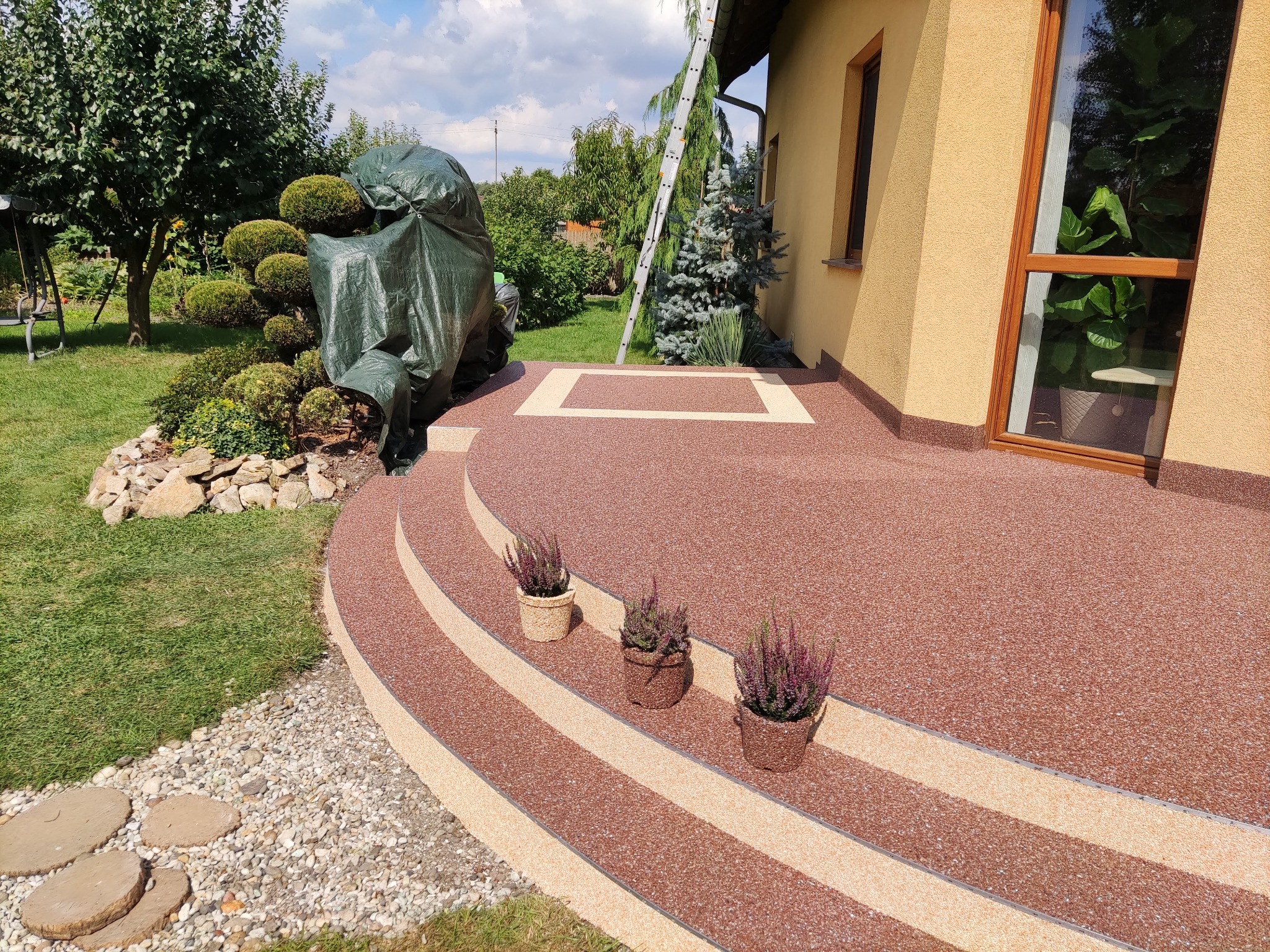 Wykończony taras z żywicy epoksydowej w odcieniach brązu i beżu, z trzema doniczkami wrzosów na stopniach, drabina oparta o ścianę domu, ogród z formowanymi krzewami w tle.