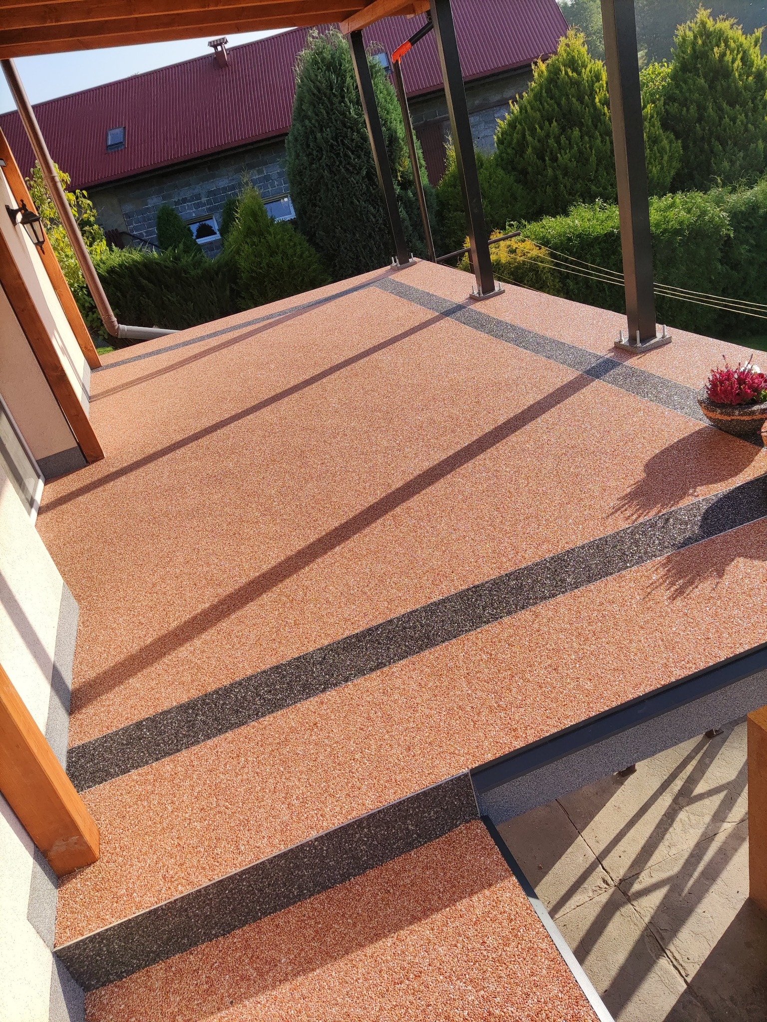 Wykończony balkon z żywiczną, pomarańczowo-szarą posadzką z dekoracyjnymi, szarymi pasami, widok z góry na zewnątrz, w tle zieleń i czerwony dach domu.