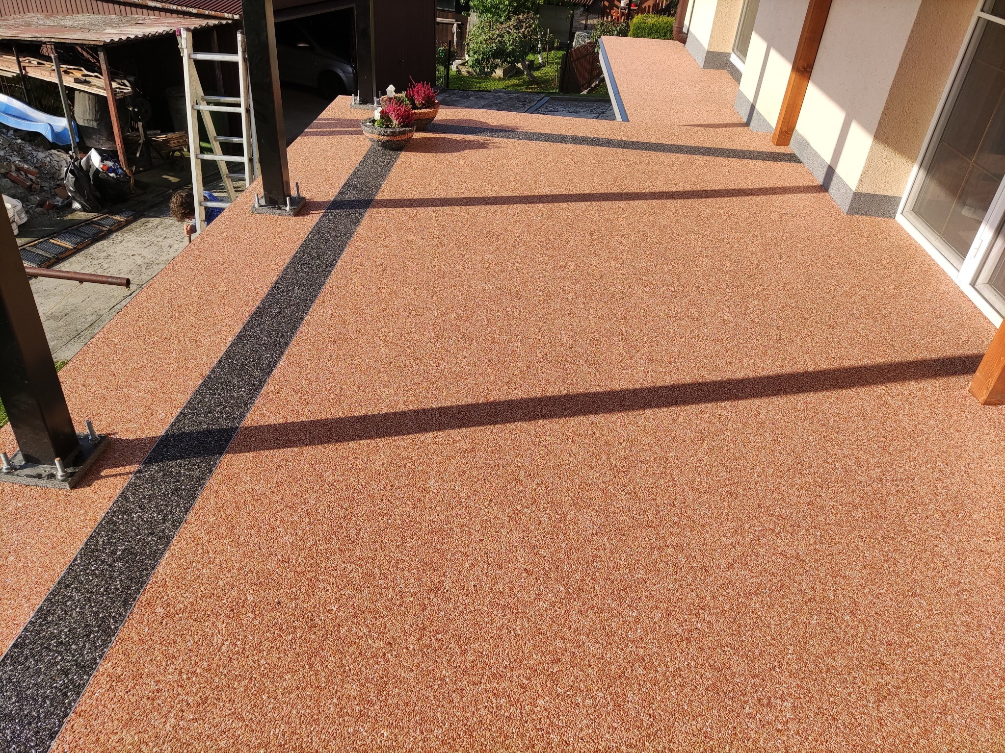 Wykończony balkon z żywicznej, pomarańczowo-brązowej posadzki z czarnym, dekoracyjnym pasem, donice z kwiatami na skraju, widoczne elementy konstrukcji dachu i drabina.