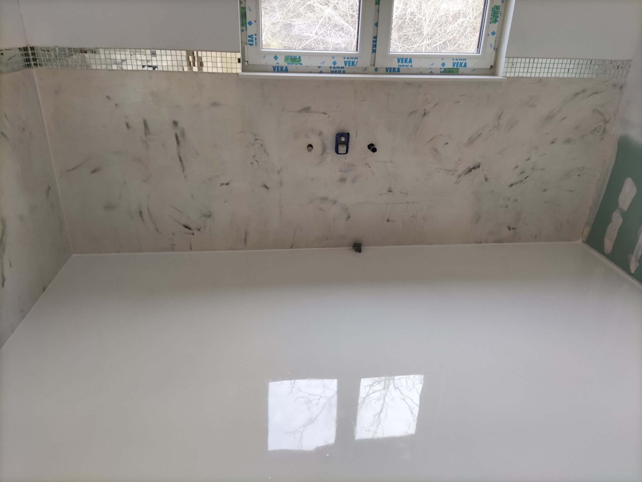 Wnętrze łazienki w trakcie remontu, ściany z efektem marmuru, fragment ściany wykończony mozaiką, widoczne instalacje elektryczne, podłoga w jasnym kolorze z odbiciem okna.