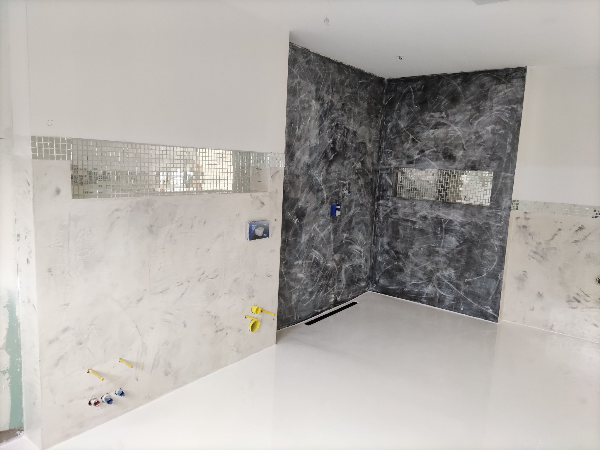 Nowoczesna łazienka w trakcie wykończenia: ściany pokryte dekoracyjną szpachlą w odcieniach szarości i bieli, mozaika lustrzana, biała posadzka żywiczna, widoczne przyłącza hydrauliczne.